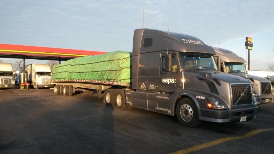A gray Volvo semi attached to a flatbed trailer with a green tarp sitting in the fuel island of a truck stop