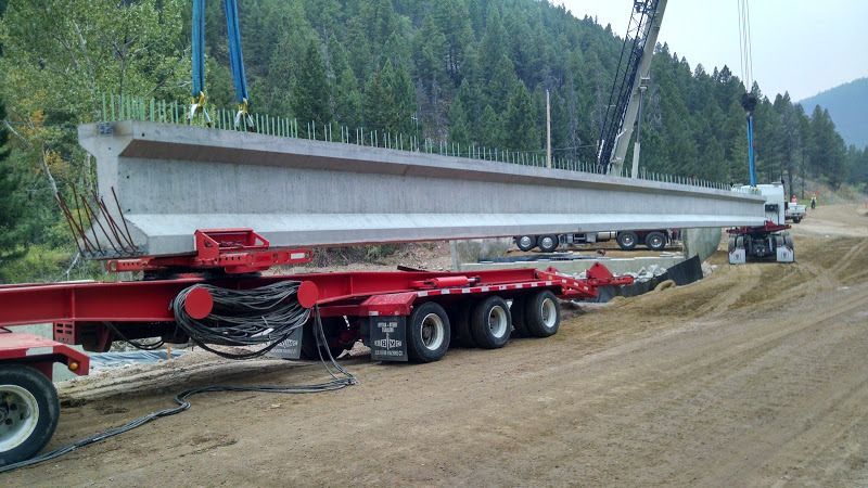 oversize flatbed trailer loaded with cement bridge beams being unloaded by a crane at construction site