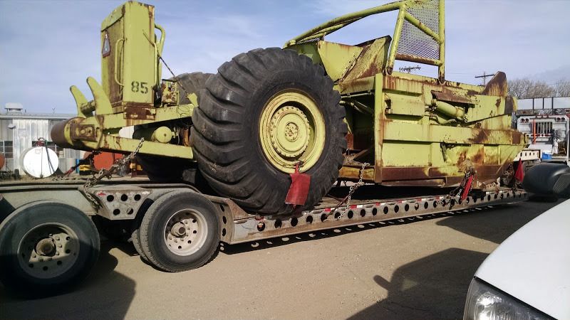 one half of green road scraper loaded and chained on flatbed trailer